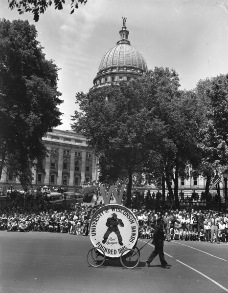Wisconsin Centennial Parade. On the street is the University of Wisconsin Bands bass drum on wheels with a depiction of Paul Bunyan on the side. In the background is a crowd lined up along the street and on the steps of the Wisconsin State Capitol.