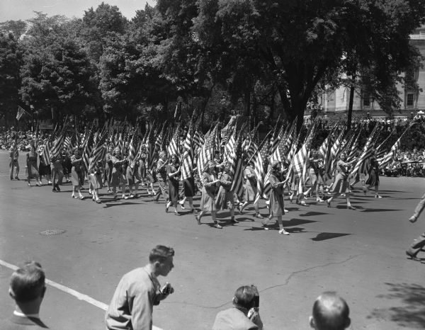 Wisconsin Centennial Parade near the Wisconsin State Capitol. A large group of Girl Scouts and Boy Scouts are marching, each with an American flag. In the foreground are two men with cameras.