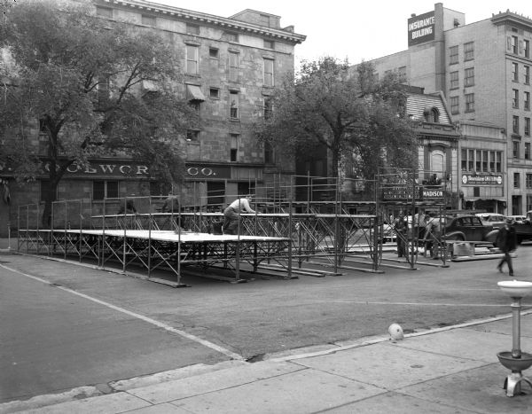 Reviewing stand for the Wisconsin centennial parade being built on Monona Avenue. Visible in the background are the Woolworth store in the Pioneer Building, 105 Monona Avenue; the Madison Theater, 111 Monona Avenue; the Davidson Cafeteria, 117 Monona Avenue, and the National Mutual Benefit Insurance Company (Beaver Building) 119-23 Monona Avenue. On the right on the sidewalk is a water fountain (bubbler).