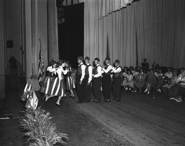 Youth in traditional Norwegian costume shown performing a folk dance on the stage of Madison West High School auditorium during the mass commencement ceremony for rural Dane County eighth grade students. The event is part of the Wisconsin centennial celebrations.