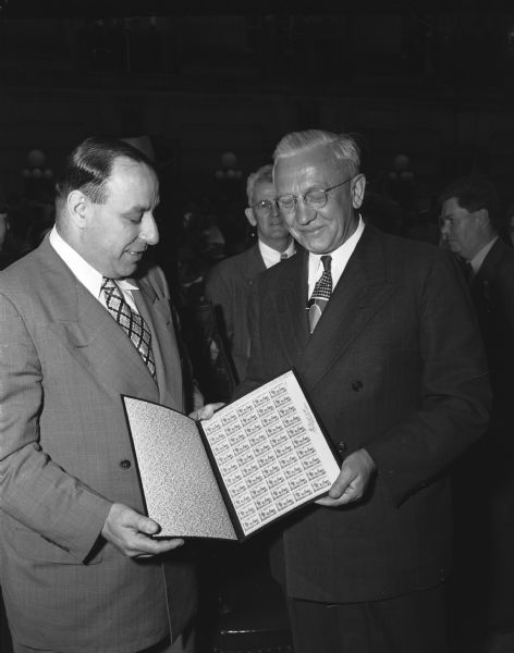 Wisconsin Governor Oscar Rennebohm shown receiving an album containing the first sheet of Wisconsin Centennial stamps from Joseph J. Lawler, third Assistant Postmaster General, as part of the state Centennial celebration.