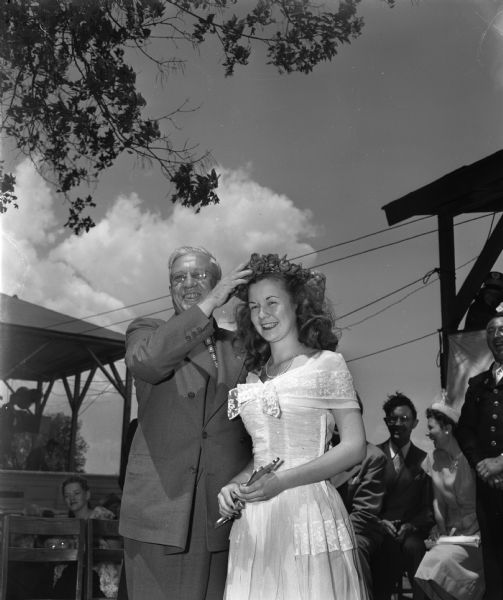 Governor Rennebohm placing a wreath of red roses on the head of Highland's Margaret McGuire, who was selected as Wisconsin's first Alice in Dairyland. Margaret holds the key to Highland as her scepter. The crowning took place in her hometown of Highland during a day-long celebration. She was also selected as queen of Wisconsin's Centennial State Fair.
