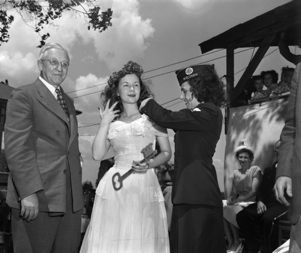 Newly selected Alice in Dairyland, Margaret McGuire, receiving an orchid from Virginia Palan, legionnaire and WAVE (Women Accepted for Volunteer Emergency Service) veteran, as governor Oscar Rennehbom looks on at the celebration of Margaret's selection in Highland. Margaret holds the key to Highland as her scepter.