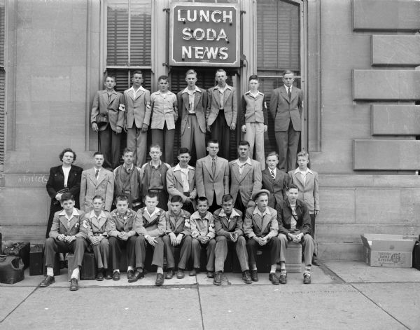 Group portrait of <i>Wisconsin State Journal</i> newspaper carriers and a few parents, taken outside the train station, who left Madison on the train for a tour of Washington, D.C. "Lunch, Soda, News" neon sign in the background.