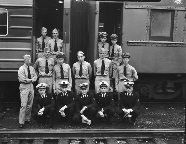 Group portrait of 16 members of the University of Wisconsin Naval Reserve Officers Training Corps in front of two railroad cars. They left for San Francisco on the first lap of a two month training cruise.