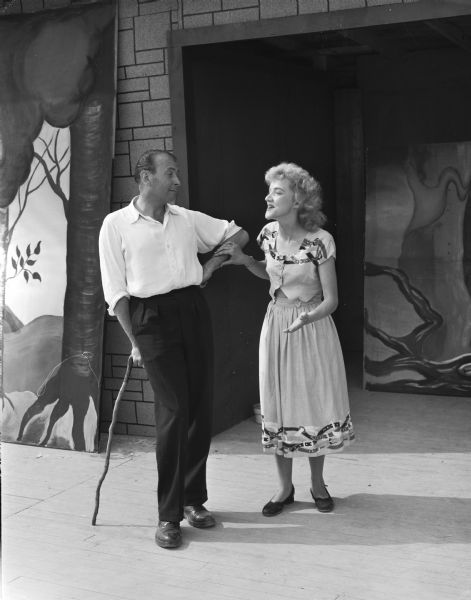 Performers Olaf Ussing, leaning on a cane, and Carole Branley rehearsing for the presentation of "The Drunkard", a Crater Players production. They are in front of a set on stage at their outdoor theater near Blue Mounds.