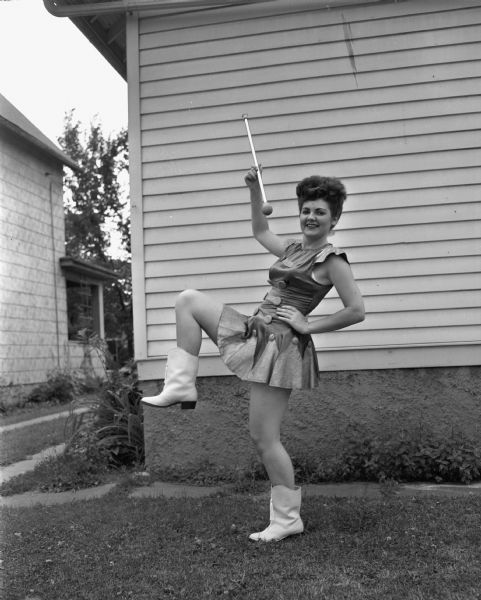 Mrs. Schafer (?) in a drum majorette costume in a marching position with a baton and wearing boots, posing outdoors near a house.
