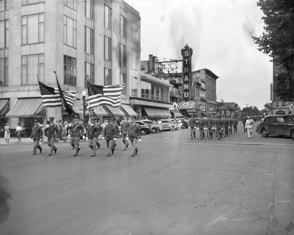 Milwaukee Aerie Eagles Conventioneers marching in their 46th annual parade, on East Mifflin Street. Manchester's store, at the corner of Wisconsin Avenue and East Mifflin Street, and the Strand Theater, is shown in the background. The flag honor guard is shown in the foreground.