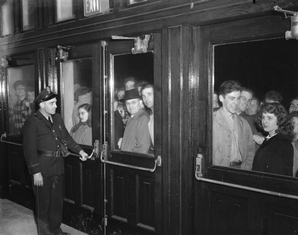 Men and women wait at the doors of the University of Wisconsin-Madison Stock Pavilion where hundreds of people had to be turned away because of lack of space when Republican presidential candidate Harold E. Stassen spoke. Shown guarding the doors is officer Joe Hammersley.