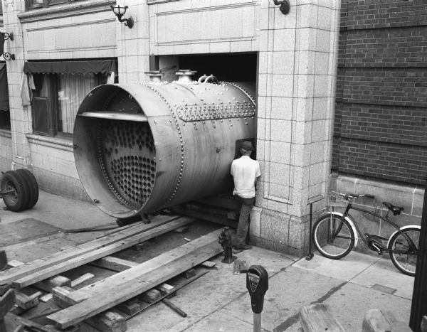 One of two new hot water boilers being moved into the basement boiler room of the Hotel Loraine. A special ramp had to be constructed into the basement, and a hole cut in the basement ceiling to get the two tanks into the boiler room. Two men are working around the boiler on the sidewalk.