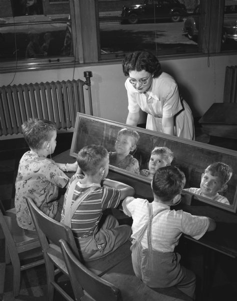 Participants in a large-scale hearing clinic held at Washington School in Madison and sponsored by the University of Wisconsin departments of speech and education with the cooperation of the state bureau for handicapped children. Three youngsters, shown wearing their new hearing aids, watch Dorothy McDaniels of Oshkosh pronounce words and then look in a mirror while imitating them.