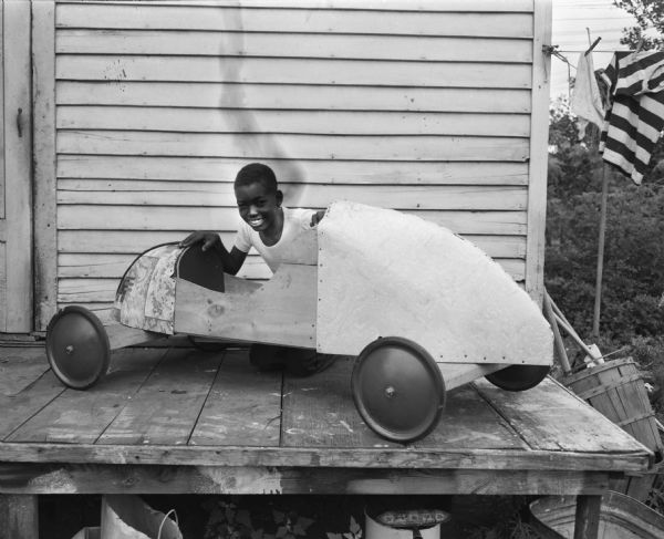 Soap Box Derby racer Virgil Johnson, posing with his car outdoors on a porch.  This image was taken before Virgil smoothed and painted his car.