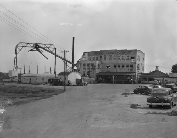 View from road of the Waunakee Canning Company building with cars in a parking lot. Men are working near the loading dock.