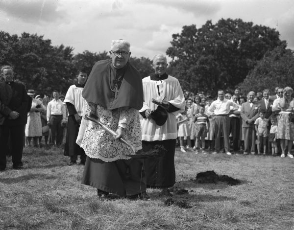 Bishop William P. O'Connor, in vestments, turning the first shovel of dirt for the building that will contain the parish auditorium and six classrooms on the first floor, and quarters for the sisters on the second floor. Looking on are Rev. Francis L. McDonnell, pastor of the parish, also in vestments, and a crowd of parishioners. The school building will be located on Mineral Point Road on a 6-acre site.