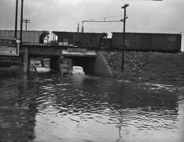 Flooded South Park Street underpass, with car stalled in three feet of water. A train is on the railroad bridge above. A young boy and girl wearing bathing suits are standing near the stalled car.