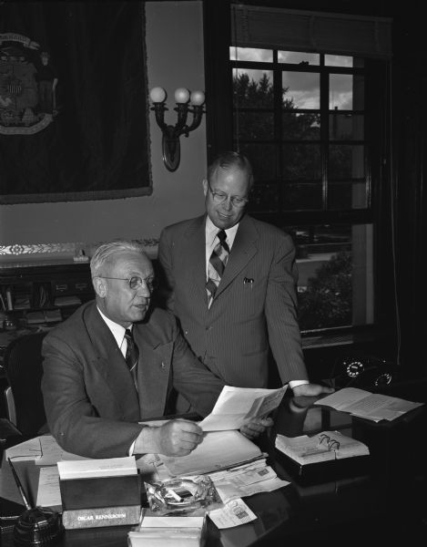 Governor Oscar Rennebohm seated at a desk at the Wisconsin State Capitol. An unidentified man is looking on.