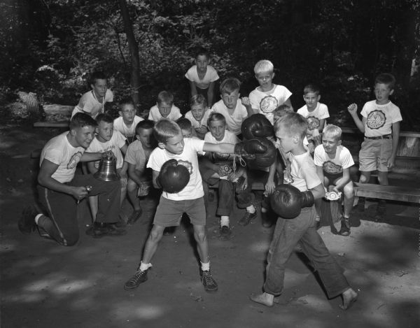 Camp Wakanda YMCA campers activities include boxing. Two boys boxing with large boxing gloves are Lawrence Fish, son of Mr. and Mrs. Leonard Fish, 842 Terry Place, and Warren Comstock, son of Mr. and Mrs. Reginald Comstock, 1034 Seminole Highway. The counselor on the left is David Brown, son of Mrs. Mildred Brown, 332 West Washington Avenue. Other campers are looking on as the boys box.
