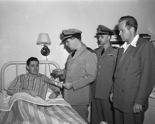 Leroy L. Dalton, (in a hosptial bed at left) shown receiving the United States Navy Air Medal for meritorious achievement during his fifth mission as a gunner or bomber during action in the Pacific in 1944. Awarding the medal are, left to right: Lieut. Commander Raymond J. McCue, Lieut. Commander G.L. Block, and John Jamieson, public relations officer of the Madison naval reserve. Mr. Dalton was discharged from the navy March 3, 1948, and was enrolled at the University of Wisconsin when he contracted tuberculosis. He is a patient at the Lake View sanatorium.