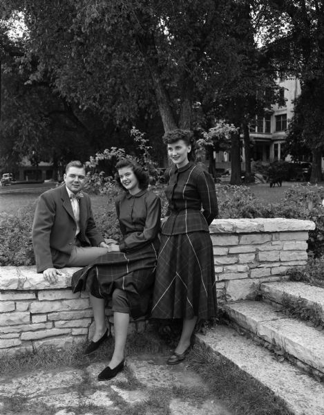 Three models are seated on the stone wall of the Memorial Union, University of Wisconsin-Madison campus, wearing clothes they will model in the up-coming style show. Left to right are: Gene McAllister, Joliet, Illinois; "Sis" McKenzie, and Florence Chudik, Milwaukee, Wisconsin.