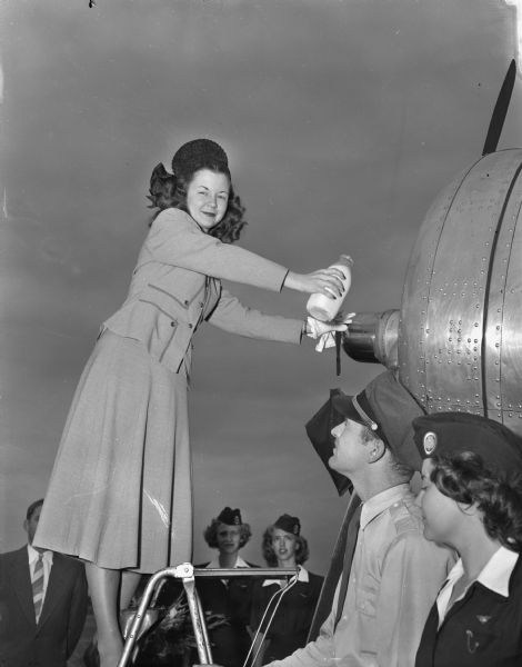 Margaret McGuire, Alice in Dairyland, at left, christens the "Northliner Alice in Dairyland" with a bottle of milk at the Madison municipal airport, with North Central Airlines pilot Robert Baker, in center, and Girl Scout Norma Trindle, at right looking on.