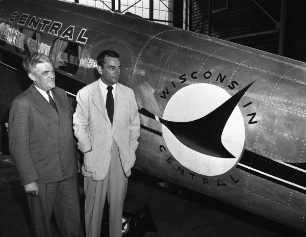 Two men standing indoors next to a Wisconsin Central Airplane.