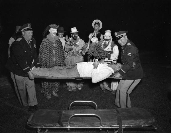 Five clowns and two police officers, with Joseph L. "Roundy" Coughlin on a stretcher, at the Knights of Columbus - Zor Shrine Softball Show at Breese Stevens Field.