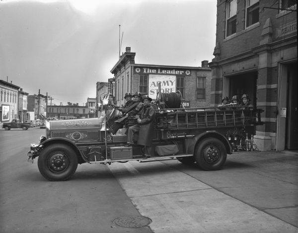 Looking south on South Webster Street with the soon to be "retired" pumper fire truck and six fire fighters, parked in front of the Central Fire Station, 18 South Webster Street. In the background is the side of the Leader Army Store, 126 East Main Street.