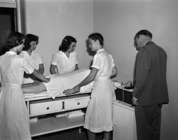 Mendota State Hospital student volunteers shown assisting Dr. Adolf Soucek give a patient electric shock treatment. The University of Wisconsin students are, left to right, Lucille Schultz from Manitowoc, Virginia Theller from Monroe, and Ann Lahey from Two Rivers. Miriam Jespersen from Sycamore, a Beloit College student, is next to the doctor. (Mendota Mental Health Institute)