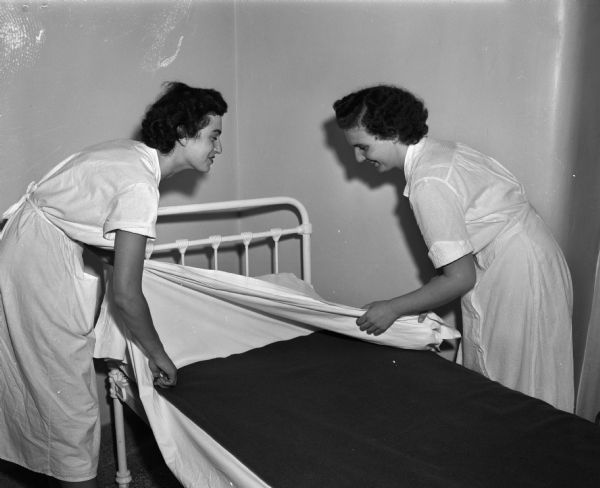 Two students making a bed at the Mendota State Hospital during their University of Wisconsin program to learn the inside workings of a mental institution. They are, from left, Eva Hirsch from Milwaukee, a Lawrence College Student, and Eleanor Wooten from Beloit, a Beloit College student. (Mendota Mental Health Institute)