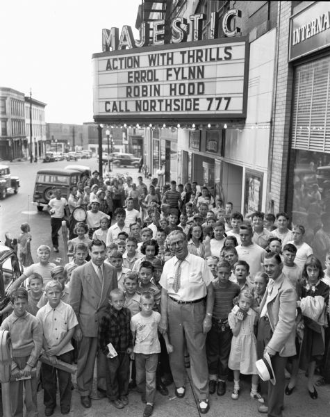 From the left: Ray Rockow, manager of the Majestic Theater on King Street; Roundy, <i>Wisconsin State Journal</i>; and Cliff Harm, Montgomery-Ward and Co. They are surrounded by a large group of children in front of the Majestic Theater. The theater marquee announces Errol Flynn in "Robin Hood."