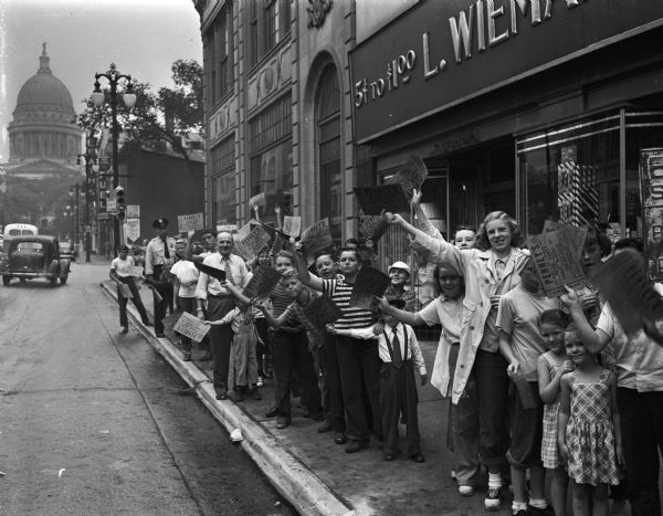 Children lined up on sidewalk in front of L. Wieman Co. Store, 205-07 State Street, to attend a free back-to-school movie party at the Capitol Theatre. In the background is Yost's Department Store and the Wisconsin State Capitol.