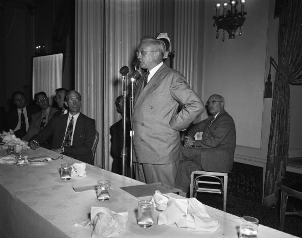Gov. Rennebolm at WKOW microphone at a table with six seated men.  Probably taken at the Dane County Republican Club gathering at the Hotel Loraine.