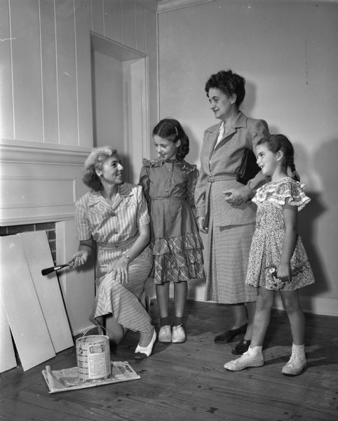 Mrs. David E. (Doris) Green and her two daughters, who have just moved into their new home at 1525 Sumac Drive, are shown being visited by Mrs. John (Nell) Hicks, member of the Shorewood Hills hospitality committee. Pictured left to right: Mrs. Green, shown painting her living room; Rowena Green, 10; Mrs. Hicks, and Pamela Green, 12.  Dr. Green is the new director of the Enzyme Institute at the University of Wisconsin, and Mrs. Green is a professional dress designer who was head of the design department of the Cambridge School of Art in England before her marriage. The Greens moved to Madison from New York City.