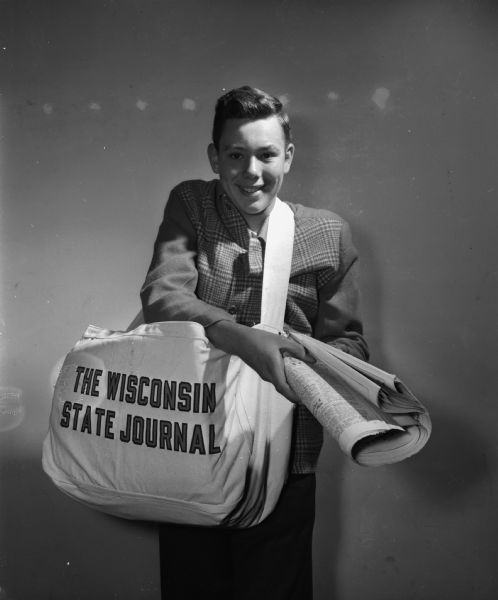 Portrait of Robert Stephen Goddertz, a <i>Wisconsin State Journal</i> newspaper delivery boy, wearing a large delivery bag and extending a newspaper toward the camera. Goddertz delivered to 108 customers on Rutledge Street for $7.00 a week. He lived at 2102 Oakridge Avenue.