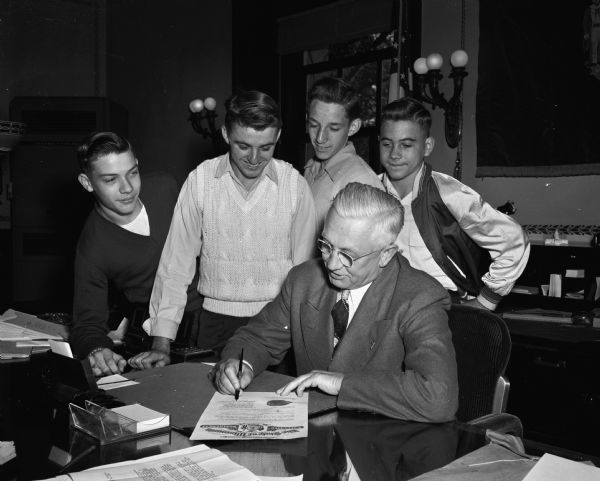 Governor Oscar Rennebohm signing a proclamation for Newspaperboy Day.  Observing the signing ceremony are four newspaper boys. From left to right are: Ken Sipes, Jr., 1027 East Johnson Street, Tim Schyska, 810 Clymer Place, Eugene Freye, 49 South Marquette, and Paul Green, 223 North Marquette.