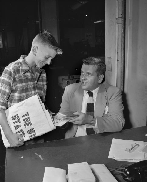 Ronnie Hinrichs, a recently hired newspaperboy for the <i>Wisconsin State Journal</i>, receiving his equipment from Howard McCaffery, city circulation manager.