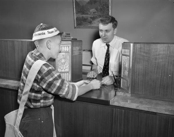 Ronnie Hinrichs, newspaperboy for the <i>Wisconsin State Journal</i>, presents his savings book to James Allaby, savings teller at the Madison Bank and Trust Co., before opening an account.