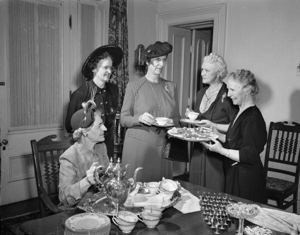 Mrs. Frank V. Powell, seated at tea table, program chairman of the convention of the Wisconsin Federation of Women's Clubs, is pouring tea for four ladies. Standing left to right: Mrs. K.L. Kleinpoll, president of the Federation; Mrs. J.L. Blair Buck, president of the General Federation of Women's Clubs; Mrs. Oscar (Mary) Rennebohm, wife of the Governor, and Mrs. L.F.(Leslie) Van Hagan, president of the Madison Women's Clubs. The tea is being held at the Governor's residence, where Mrs. Rennebohm is hosting the event.
