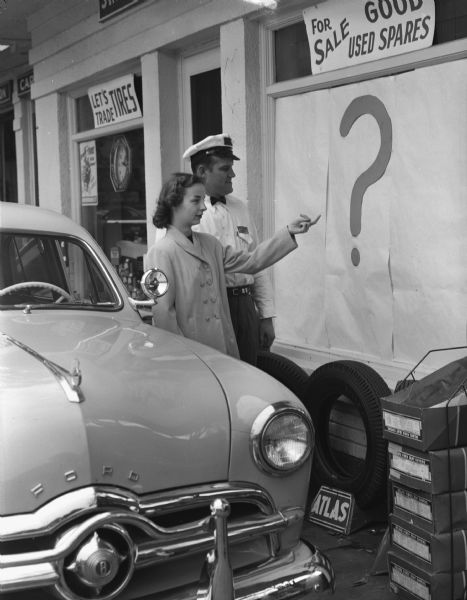 A woman standing between a new Ford automobile and a capped and uniformed service technician pointing at a question mark covered window.