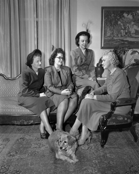 Group portrait of three members of the scholarship committee of the Madison Business and Profession Women's Club meeting at the residence of the governor with Mrs. Oscar (Mary) Rennebohm, wife of the governor. A dog sits on the floor at Mrs. Rennebohm's feet. Pictured left to right: Miss Vivian Addison, Southern Wisconsin Produce Company; Miss Ruth Hull, Bowman Dairy; Miss Louella Loken, head surgical nurse at Madison General Hospital, and Mrs. Rennebohm, who will be hosting an evening fundraising party for the scholarship fund in the near future. Miss Hull won the scholarship in 1947 and is now an active member of the club.
