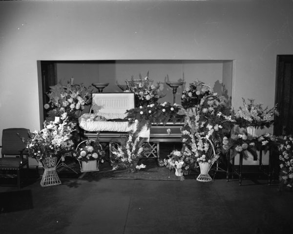 Open casket containing body of a man, and large floral displays surrounding it.