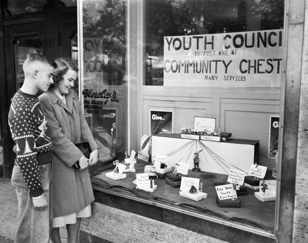 James Ketchum, left, and Annette Nelson, right, members of the Madison Youth Council, admire their display in the window of Moseley's Book Store, which was arranged by the Youth Council to promote the current Community Chest Drive. James is the son of Mr. and Mrs. L.W.(Myrtle) Ketchem, 1014 Tumalo Trail, and Annette's parents are Mr. and Mrs. Robert A. (Esther) Nelson, 1005 Oakland Avenue.