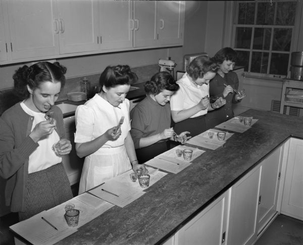 Four University of Wisconsin home economics students taste testing food samples in a home economics laboratory. Left to right: Louise Page, Payson, Utah; Mrs. Hazel (Jobelle) Shands, Eagle Heights; Margaret Wallace, Dearborn, Michigan; Engeberg Virge, 661 Mendota Court; and Enas Hansen, Wichita, Kansas.