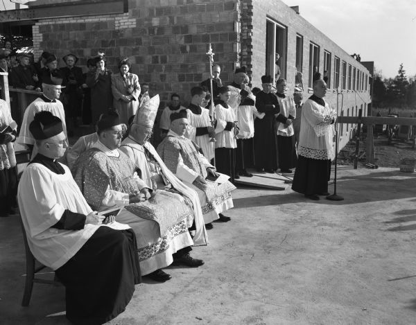 Cornerstone laying ceremony at the new Queen of Apostles seminary, 5810 Cottage Grove Road. Seated, left to right, are Rev. Edward Kinney, Madison, secretary to the bishop; Rev. Harold Liebl, Pallottine college, Milwaukee, a deacon of honor to the bishop; Bishop William P. O'Connor; and Rev. Christopher Meyer, Pallottine College and also a deacon of honor. At the microphone to the right is the Very Rev. Joseph De Maria, Milwaukee, provincial of the American Province of Pallottine fathers.