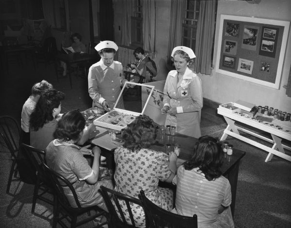 Gray Ladies of the Dane County Red Cross teaching jewelry-making to a group of patients at the Mendota hospital for the mentally ill. Gray Ladies, shown at the top of the picture, are, at left, Mrs. L. Stanley Meek, and at right, Mrs. W.G. Coles.