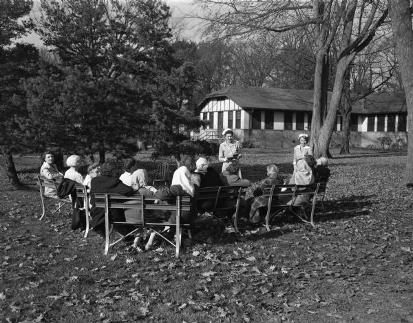Gray Ladies of the Dane County Red Cross reading outdoors to patients of the Mendota hospital for the mentally ill. Shown standing, at left, is Mrs. William Walker, and at right, Mrs. E.J. Sawyer.