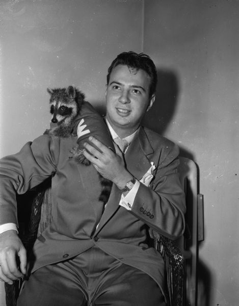 Image of George Holmes, the University of Wisconsin student Homecoming Dance chairman and raccoon keeper, seated with a young raccoon named "Regdab" (badger spelled backwards) wearing a tiny UW sweater on his shoulder.