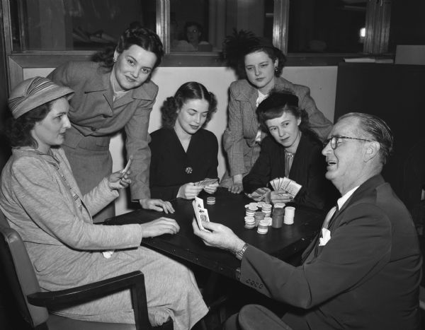 Roundy Coughlin holding playing cards with many chips in front of him at a table with five members of the Madison Junior Chamber of Commerce Auxiliary (Jaycettes). "The profits (from their fundraiser) will go into a fund being raised by the Jaycettes for the construction of a hospital for (handicapped) spastics at Camp Wawbeek near Wisconsin Dells". L to R: Bernice Johnson, kitchen committee; Betty Krohn, publicity; Ruth Prafke, decorations; Mrs. Harry Stoll, general chairman; Ruby Zein, tickets.
