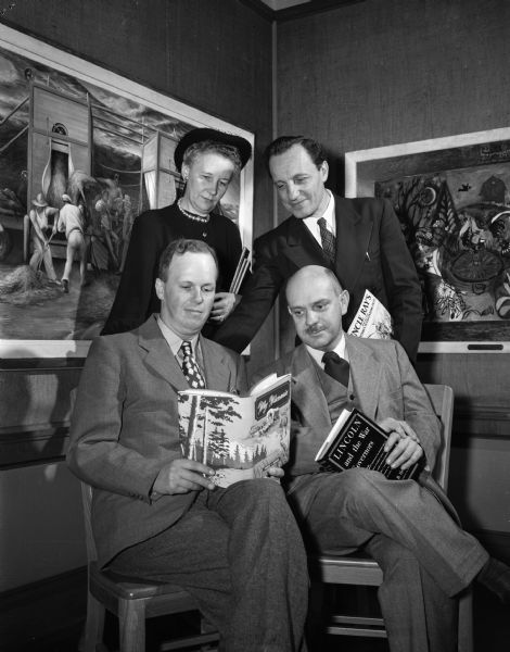 Four Madison authors are shown together holding books they have written. Seated are Francis F. Bowman and Prof. William Hesseltine.  Standing are Miss Emilie Weidenbeck and Raymon Coffman. Behind them on the wall hang two paintings.