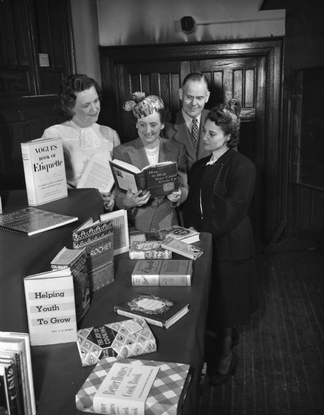 Four parents are shown admiring twelve books. They are celebrating National Book Week which is co-sponsored by their group, the Madison Council of Parents and Teachers. They are Violet Anderson, Flora Steul, Howard Van Wagenen, and Anne Bruno.